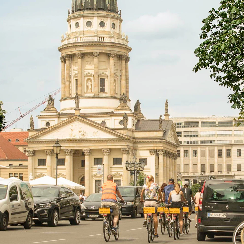 Group of people cycling on a city street with an ornate historic building and parked cars in the background.