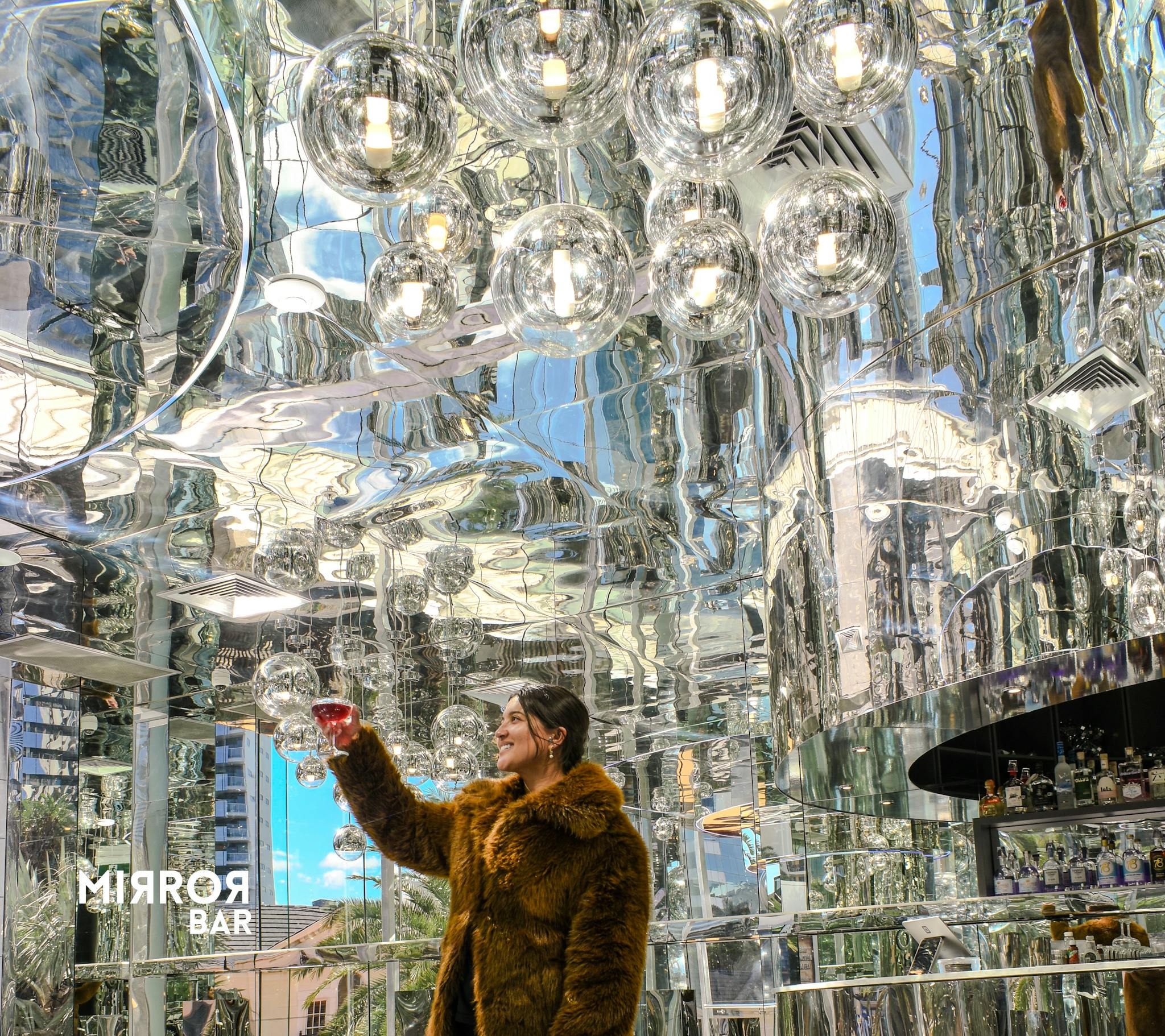 Μια γυναίκα με γούνινο παλτό κρατά ένα ποτό σε μια έντονα φωτισμένη, ανακλαστική μπάρα με γυάλινα φωτιστικά και καθρέφτες.