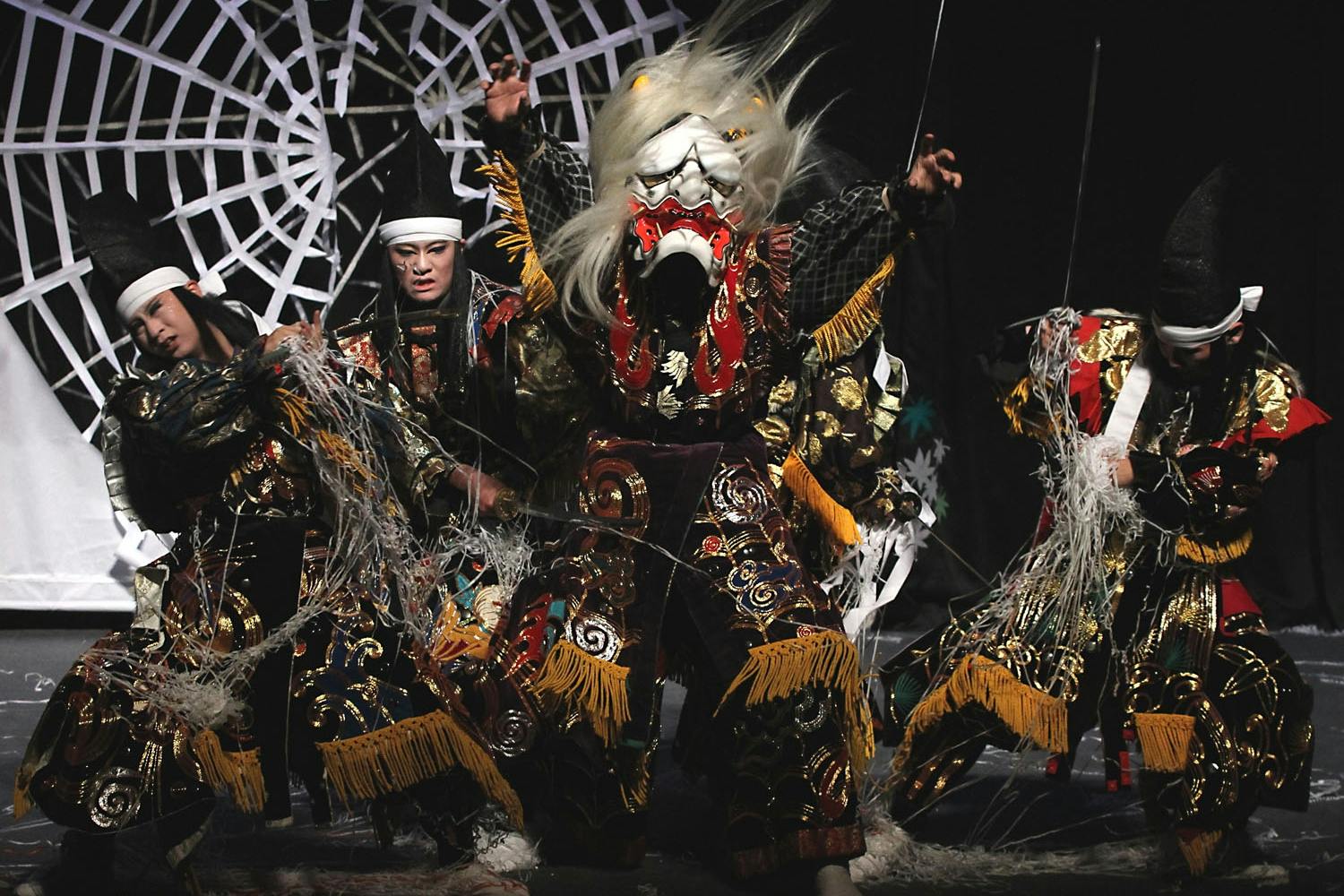 Three performers in elaborate traditional costumes and masks enact a dramatic scene in front of a spider web backdrop.