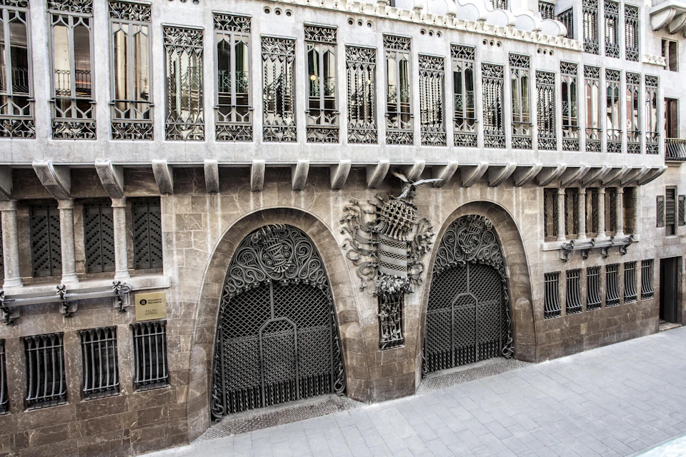 Ornate building facade with decorative ironwork and stone. Arched doorway and intricate details above windows. Sign reads "Gaudí."