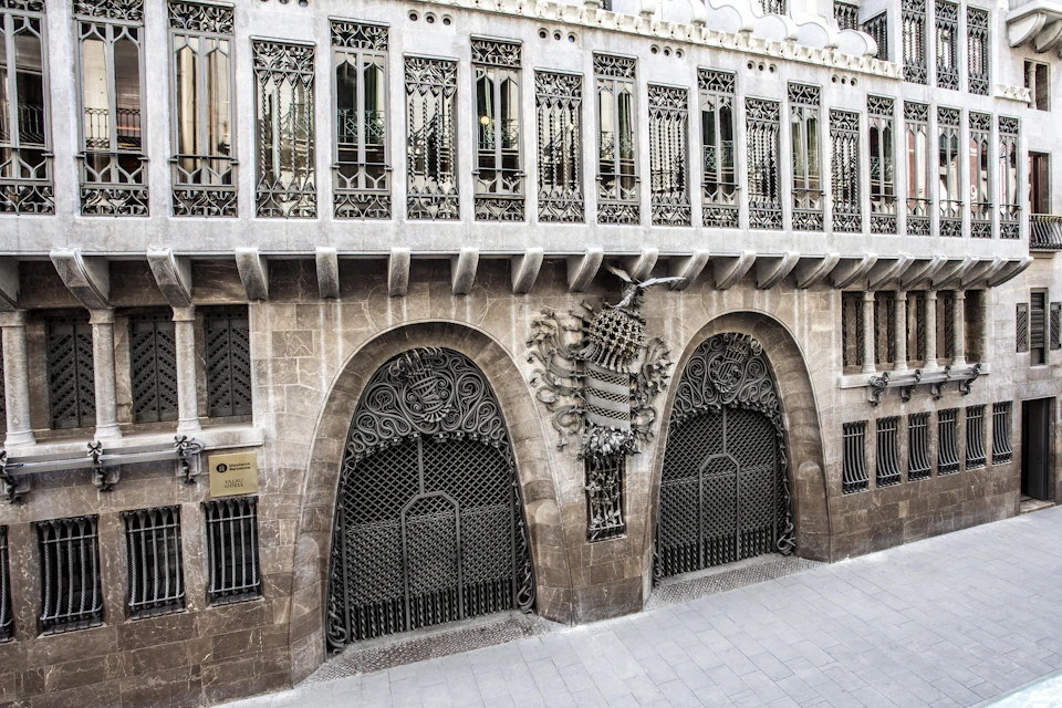 Ornate building facade with decorative ironwork and stone. Arched doorway and intricate details above windows. Sign reads "Gaudí."