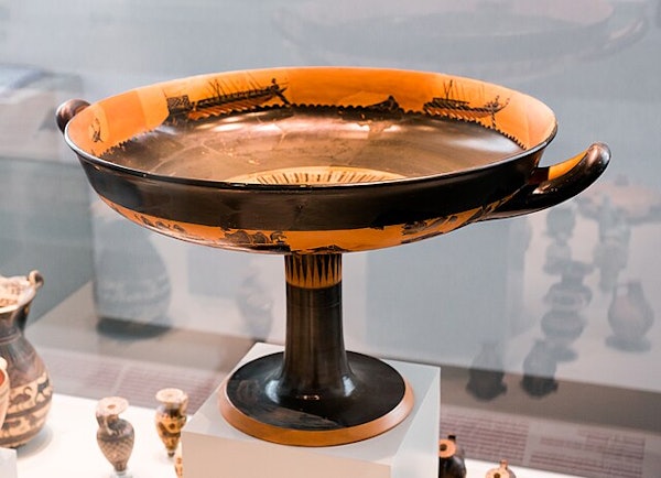A large, ancient Greek kylix with black-figure ship illustrations, displayed on a pedestal in a museum.