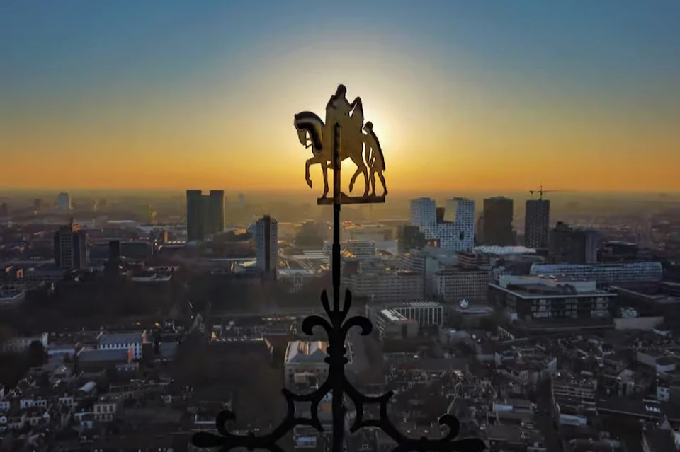Silhouette of a horse and rider atop a decorative spire, with a cityscape and sunset in the background.