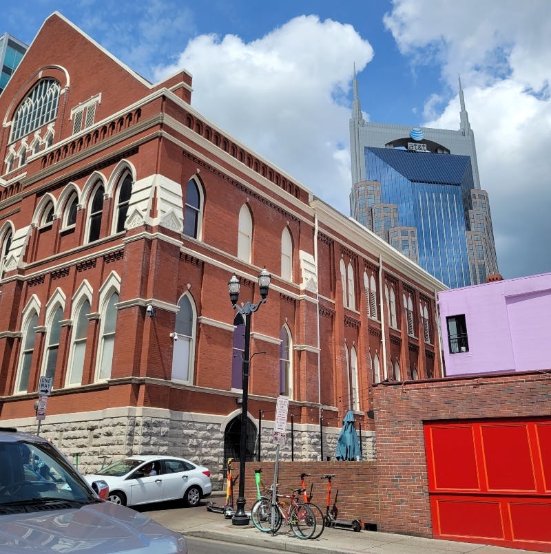 Un edificio histórico de ladrillo con ventanas arqueadas, un rascacielos moderno al fondo, coches aparcados y bicicletas en la calle.