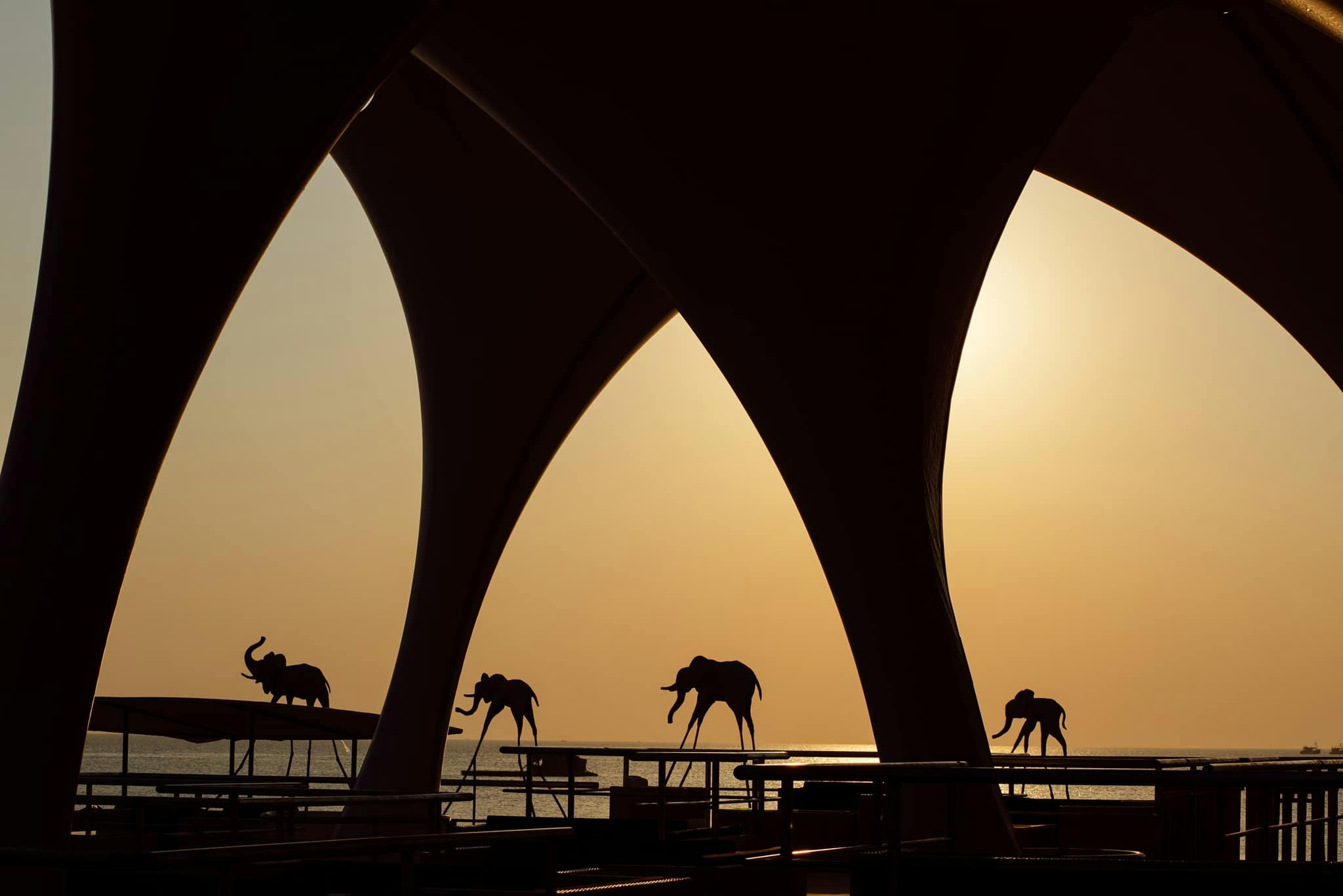 Silhouettes of elephant statues on a waterfront during sunset, framed by large architectural arches.