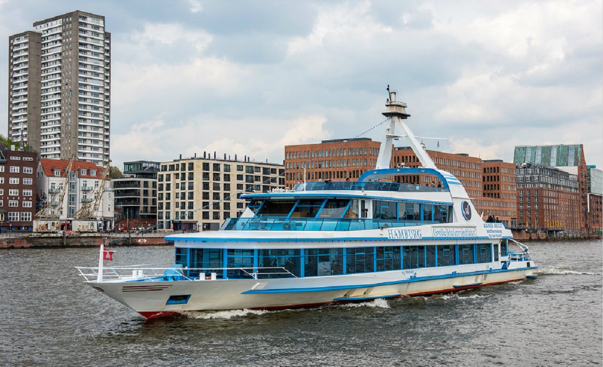 Eine blau-weiße Passagierfähre namens "HAMBURG" fährt auf einem Fluss mit modernen Gebäuden im Hintergrund.