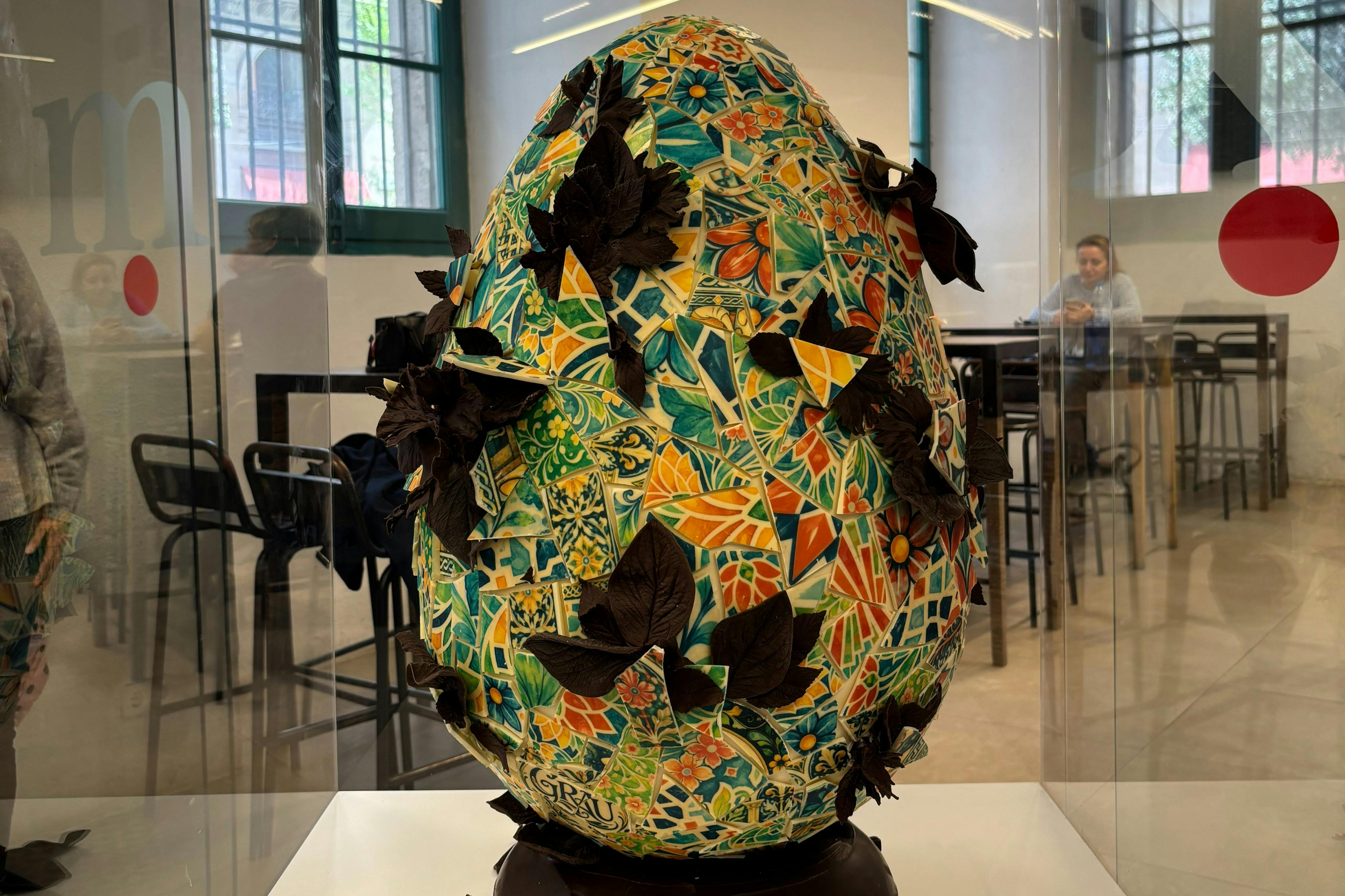 Sculpture d'œuf en mosaïque colorée avec des décorations en forme de feuilles, présentée à l'intérieur, avec un homme assis à une table à l'arrière-plan.
