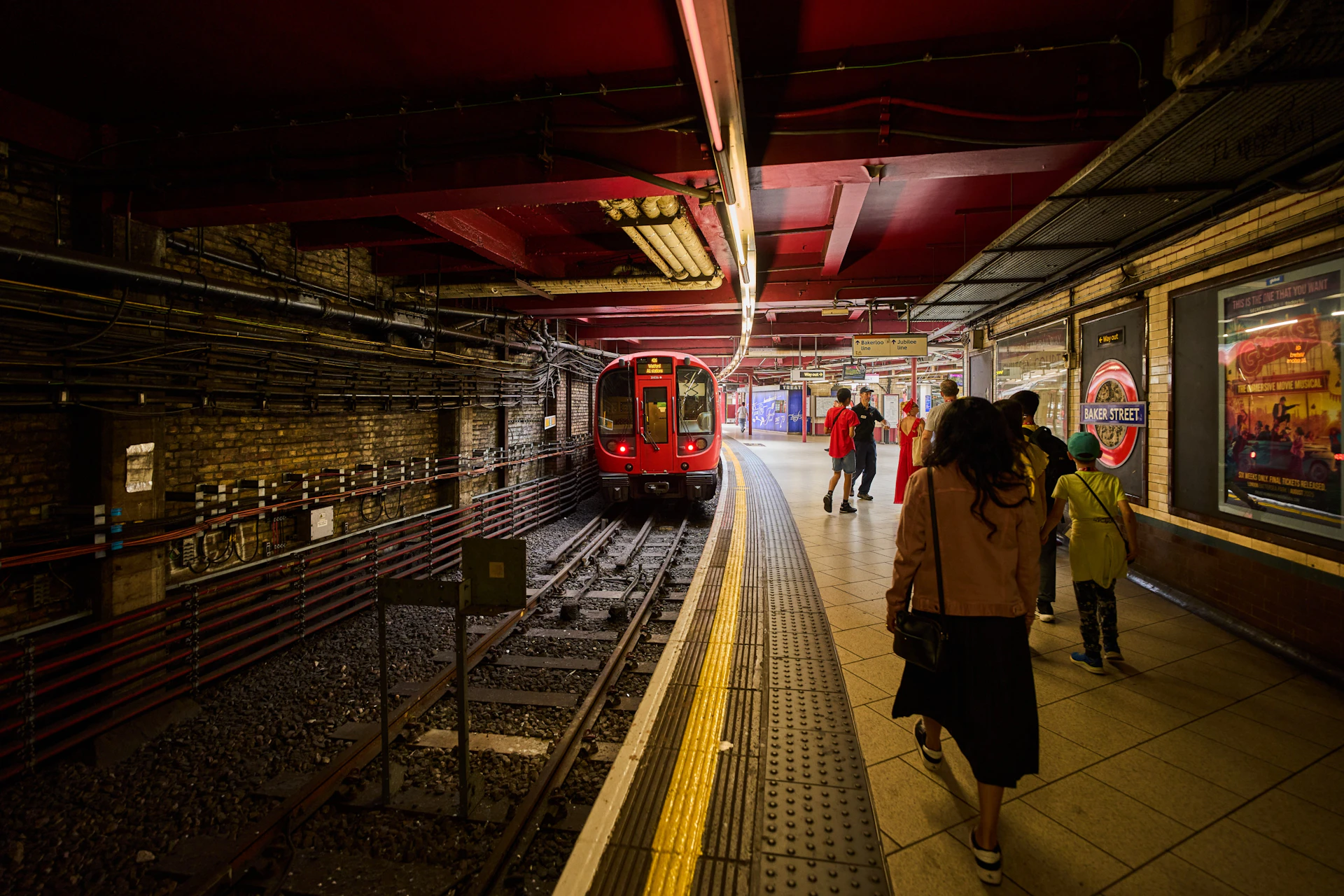 A red train stopped at an underground station platform with people walking and waiting nearby.