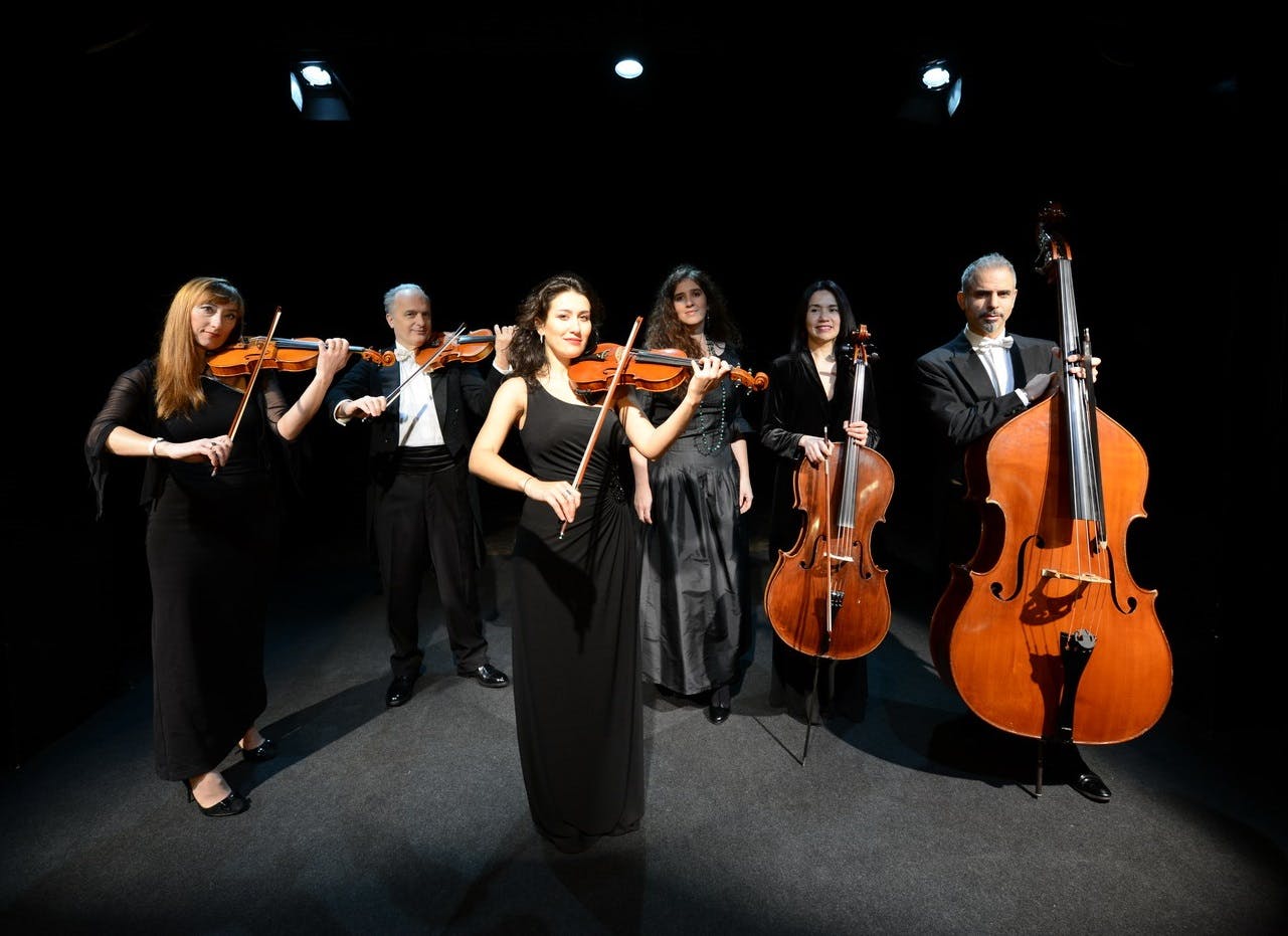 A group of six musicians in formal attire with violins, cellos, and a double bass, standing in a dimly lit performance space.
