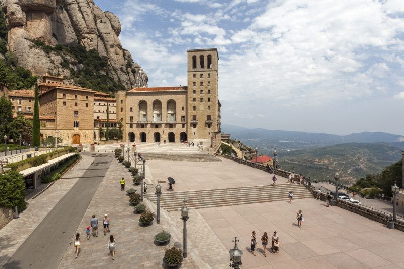 Stone plaza with scattered people near a historic building with a tall tower; rocky hillside and distant landscape under a cloudy sky.