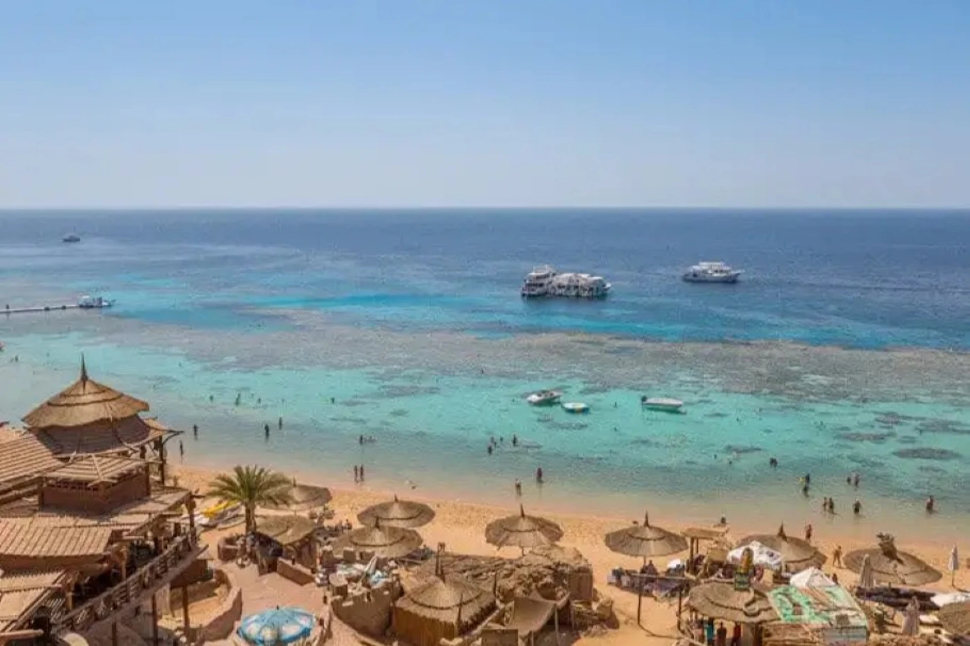Sandy beach with straw umbrellas, people in the water, boats floating in the blue sea, and a clear sky overhead.