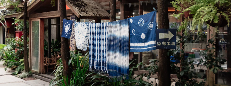 Blue and white tie-dyed fabric pieces hanging on a line outdoors near plants and a rustic building.
