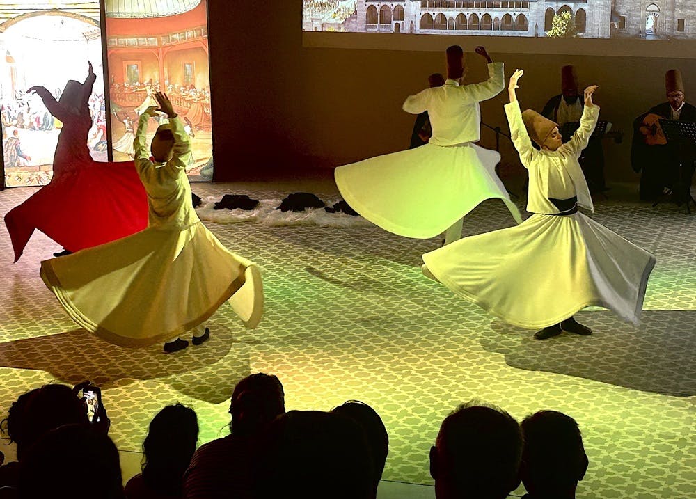 Περιστρεφόμενοι δερβίσηδες με λευκές ρόμπες χορεύουν σε ένα δάπεδο με μοτίβα, με το κοινό να κάθεται στο προσκήνιο.