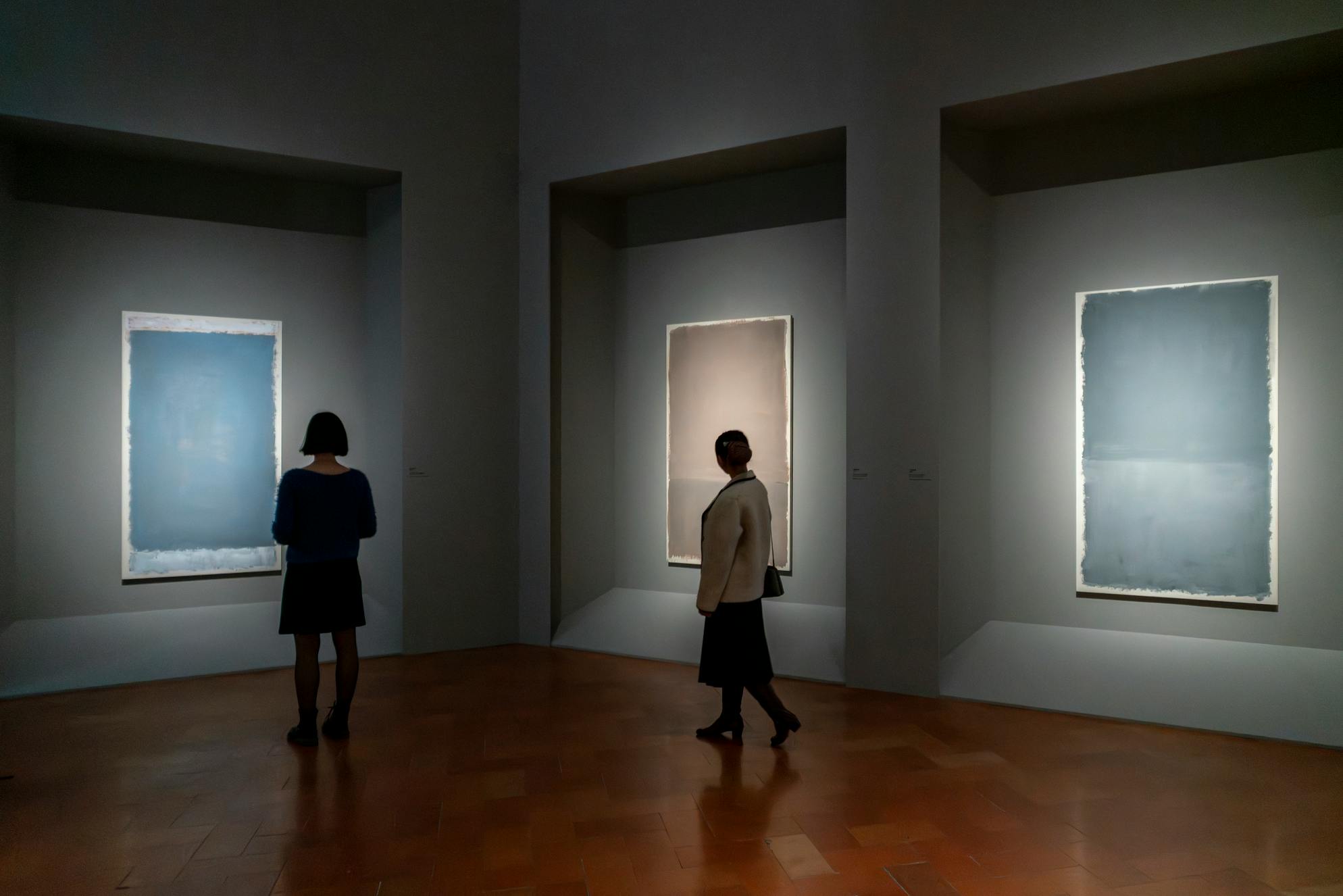 薄暗いギャラリーに展示されている抽象芸術作品を鑑賞している2人の人物。壁には額装された作品が照らされています。