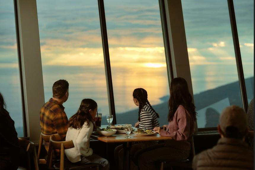 A group of four people dining at a table by a large window, overlooking a scenic sunset view.