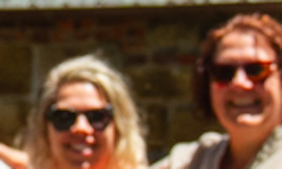 Four people smiling and posing arm-in-arm in front of a stone building with a tiled roof on a sunny day.