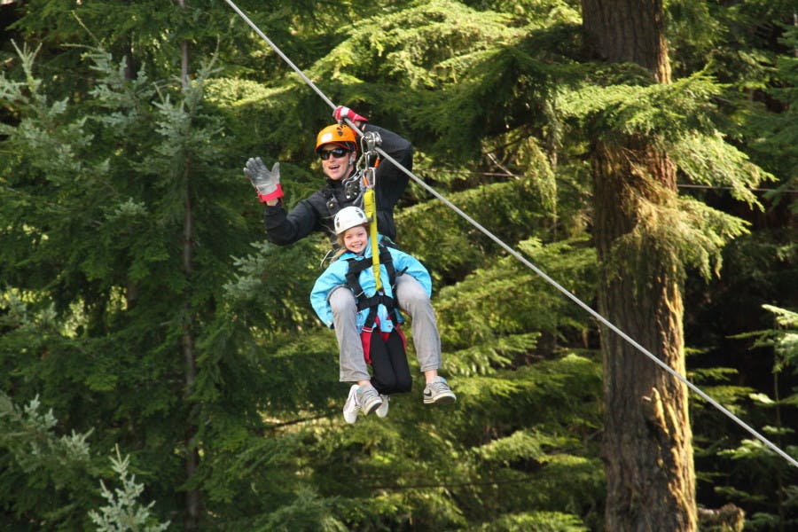 헬멧과 안전 장비를 착용하고 집라인으로 숲을 가로지르는 두 사람. 아이가 웃는 동안 어른이 손을 흔든다.