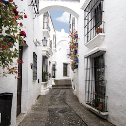 Smal kullerstensgata mellan vita byggnader med gallerfönster och färgglada blomlådor, som leder till en trappa under en valvgång.