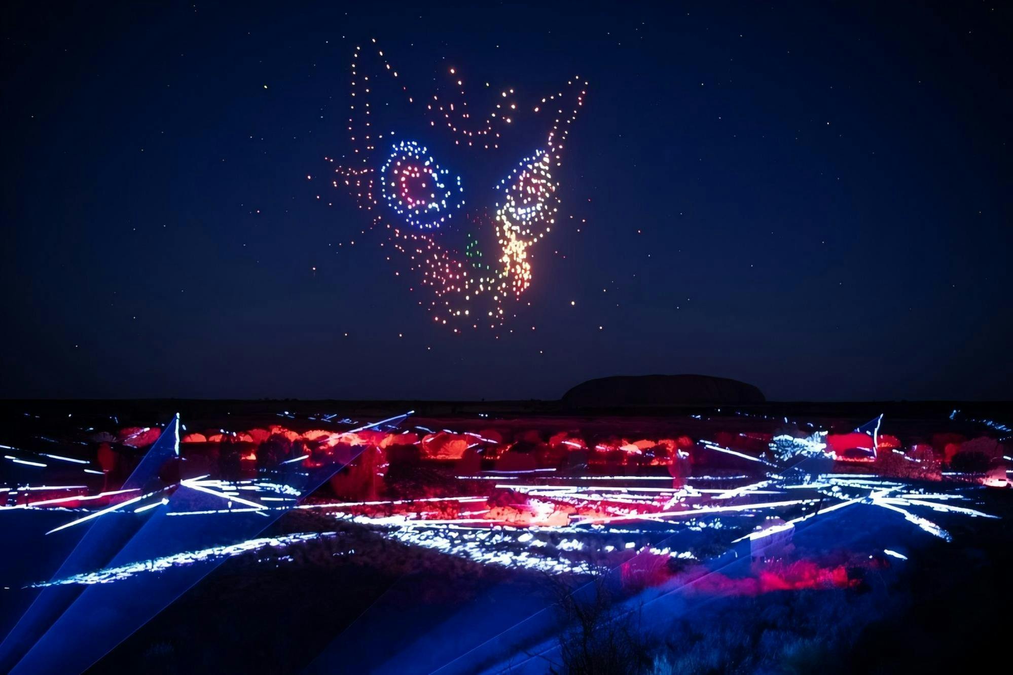 A night sky with colorful drone lights forming a fox's face. Ground below illuminated with red and blue lights.