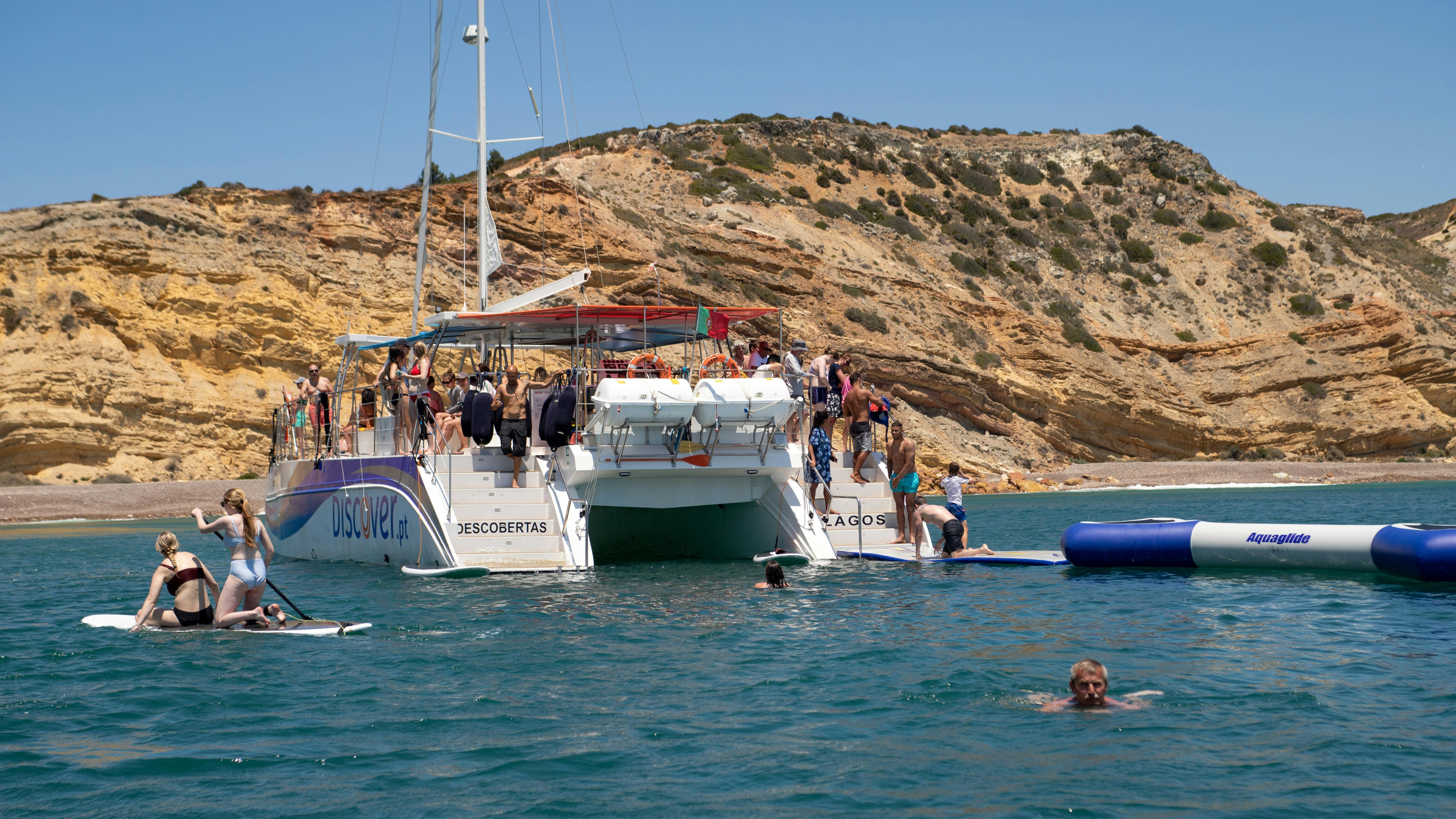 岩だらけの海岸線に近い大型のカタマランボートに乗っている人々。 デッキの上にいる人もいれば、泳いでいる人や、浮かぶプラットフォームの上に立っている人もいます。