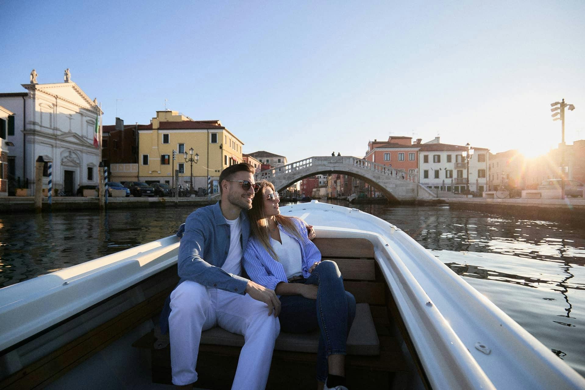 A couple seated on a boat ride, enjoying the view of a canal with colorful buildings and a bridge in the background.