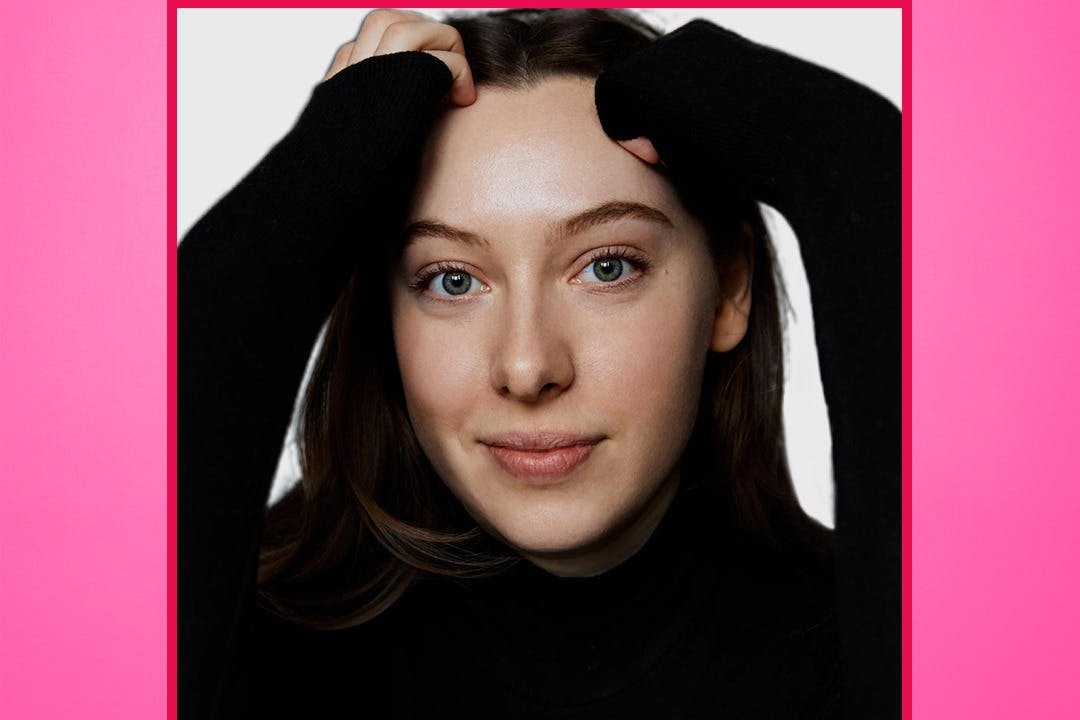 Ένα άτομο με μακριά μαλλιά που φοράει μαύρο ζιβάγκο και χαμογελάει με τα χέρια ακουμπισμένα στο κεφάλι του σε ένα απλό λευκό φόντο.