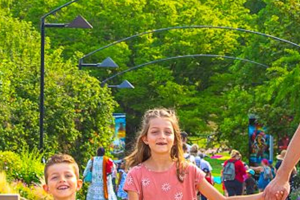 A family of four holding hands, walking under a canopy of colorful umbrellas in a park. Other people are in the background.