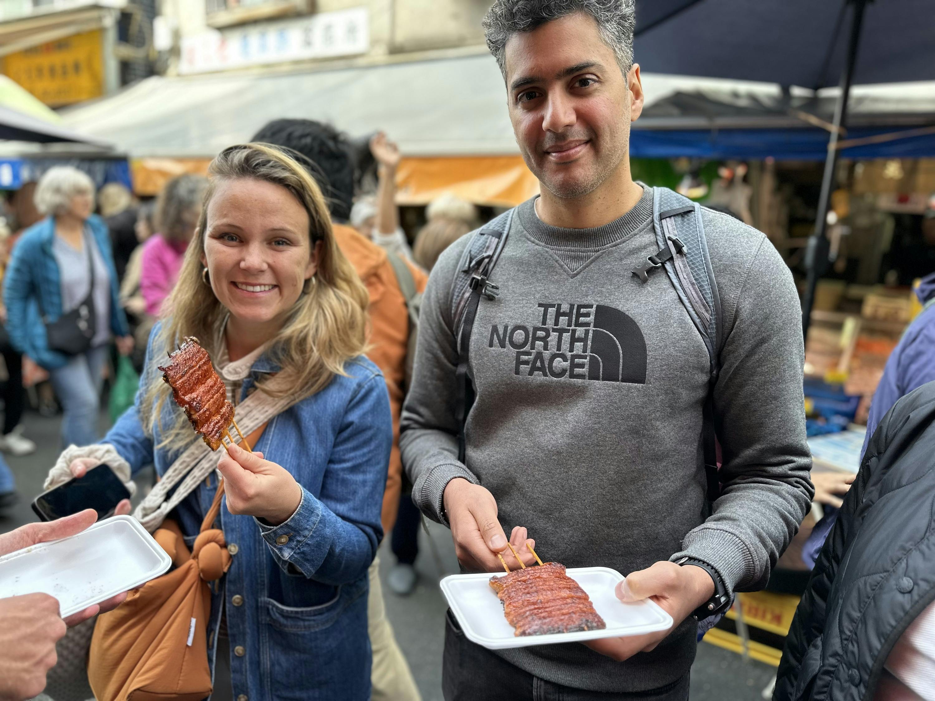 Two people holding skewers of grilled food in a busy outdoor market setting, smiling at the camera.