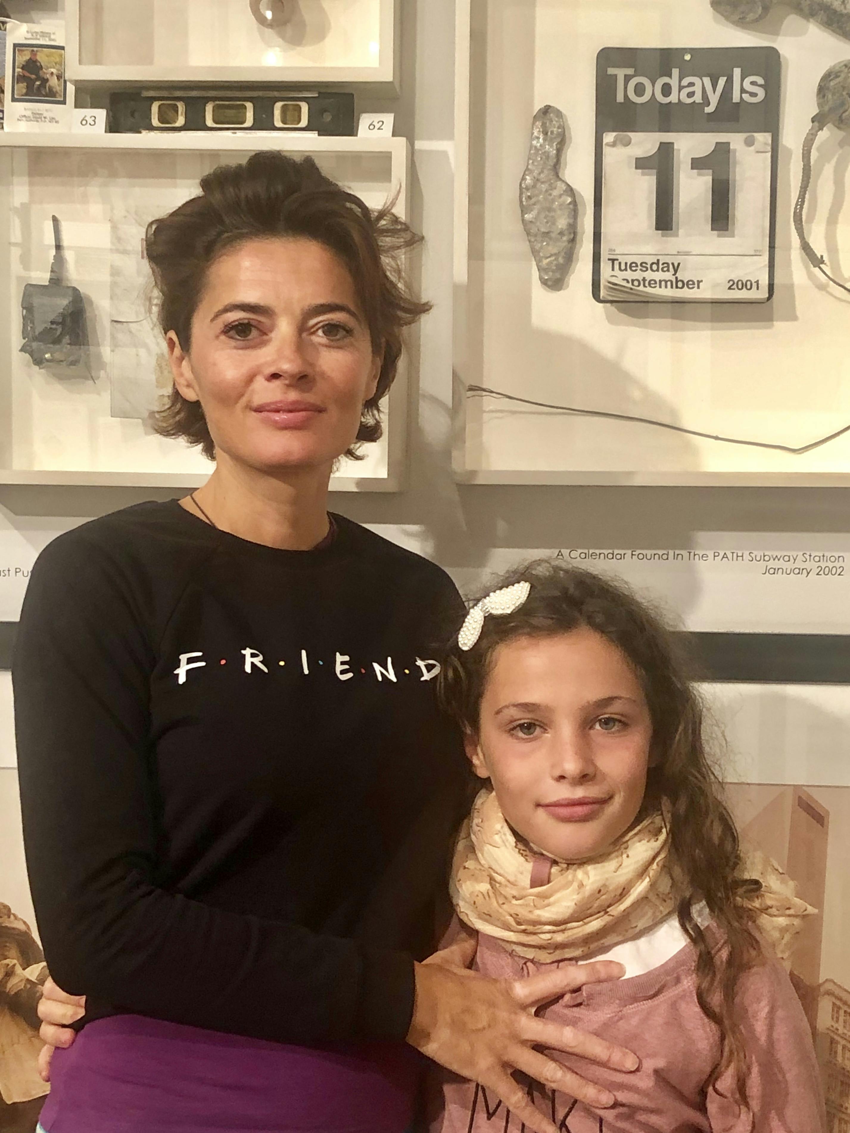A woman and a child standing indoors, with the woman wearing a black shirt that says "FRIENDS" and the child wearing a scarf.