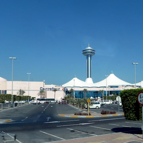 Ein Einkaufszentrum mit "Carrefour"- und "Marina Mall"-Schildern, einem hohen Aussichtsturm, einem Parkplatz, Palmen und einem Schild mit einer Geschwindigkeitsbegrenzung.