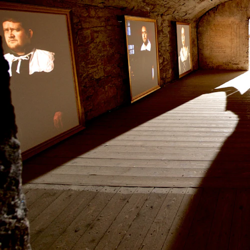 En person i tidstypisk dräkt står mot en upplyst dörröppning i ett svagt upplyst stenrum, med stora målade porträtt på väggarna.