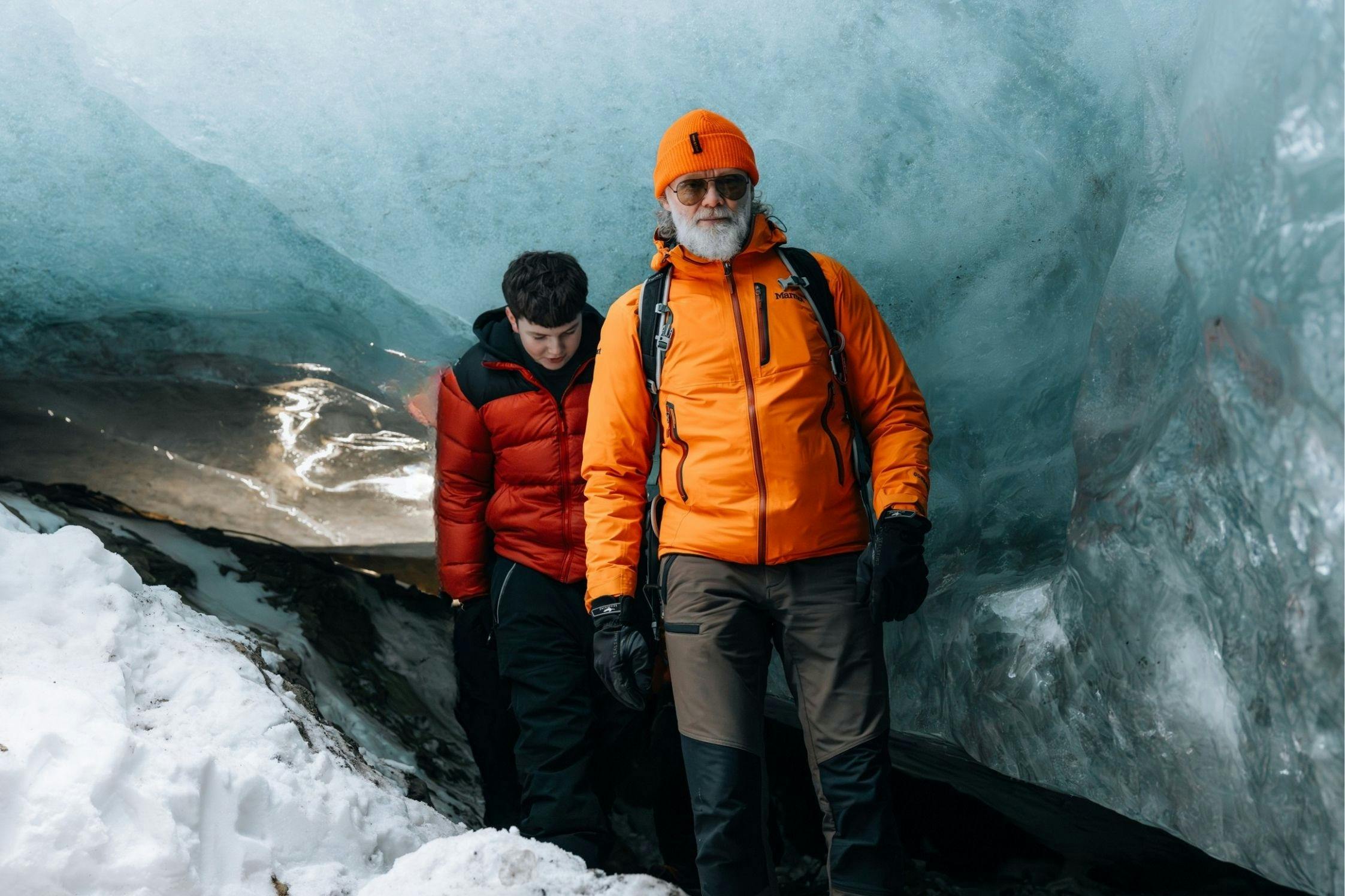 Dva lidé v zimním oblečení stojící u zasněžené ledové jeskyně, jeden v oranžové bundě a druhý v červené bundě.