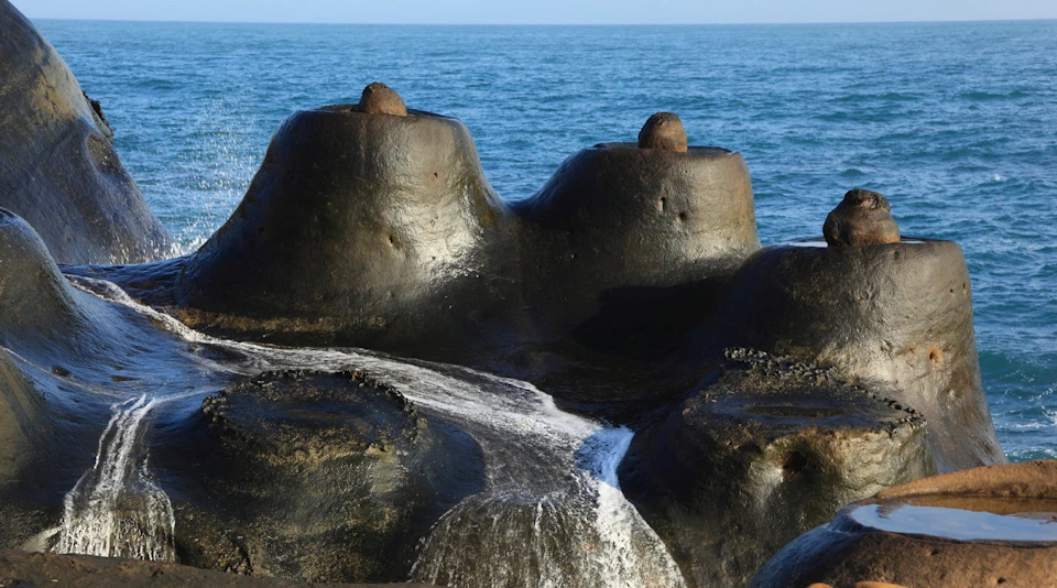 Rounded black rocks with a stream of water flowing over them, set against a blue ocean.