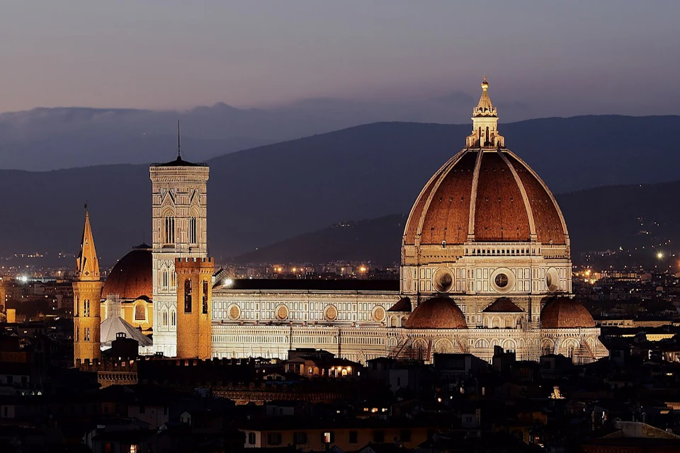 Florence Cathedral with its dome and bell tower illuminated at dusk, with a mountainous background.