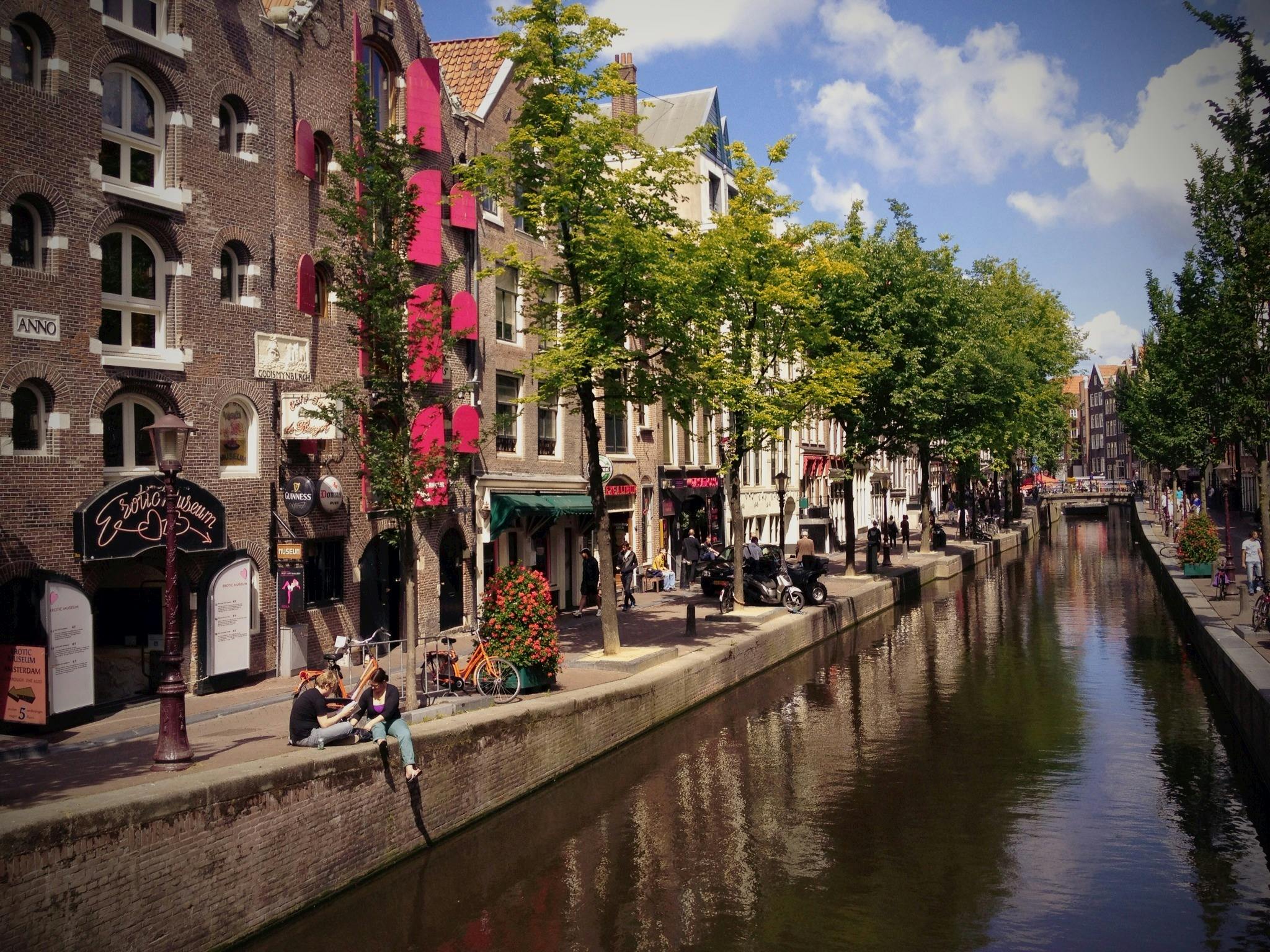 사람들은 나무와 유서 깊은 건물이 늘어선 운하 옆에 앉아 푸른 하늘 아래 야외 카페, 상점가, 주차된 자전거를 선보입니다.