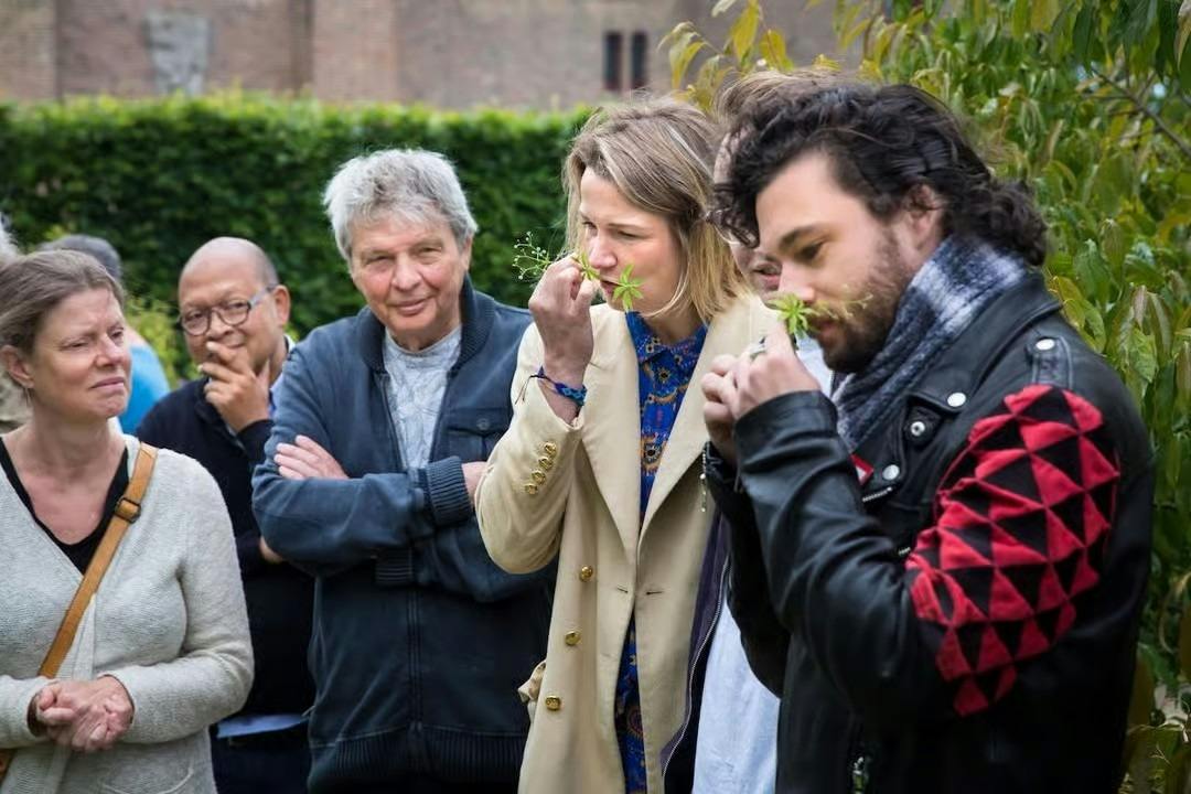 Un gruppo di persone all’aperto, alcune delle quali annusano delle piante, con del verde e un edificio in mattoni sullo sfondo.