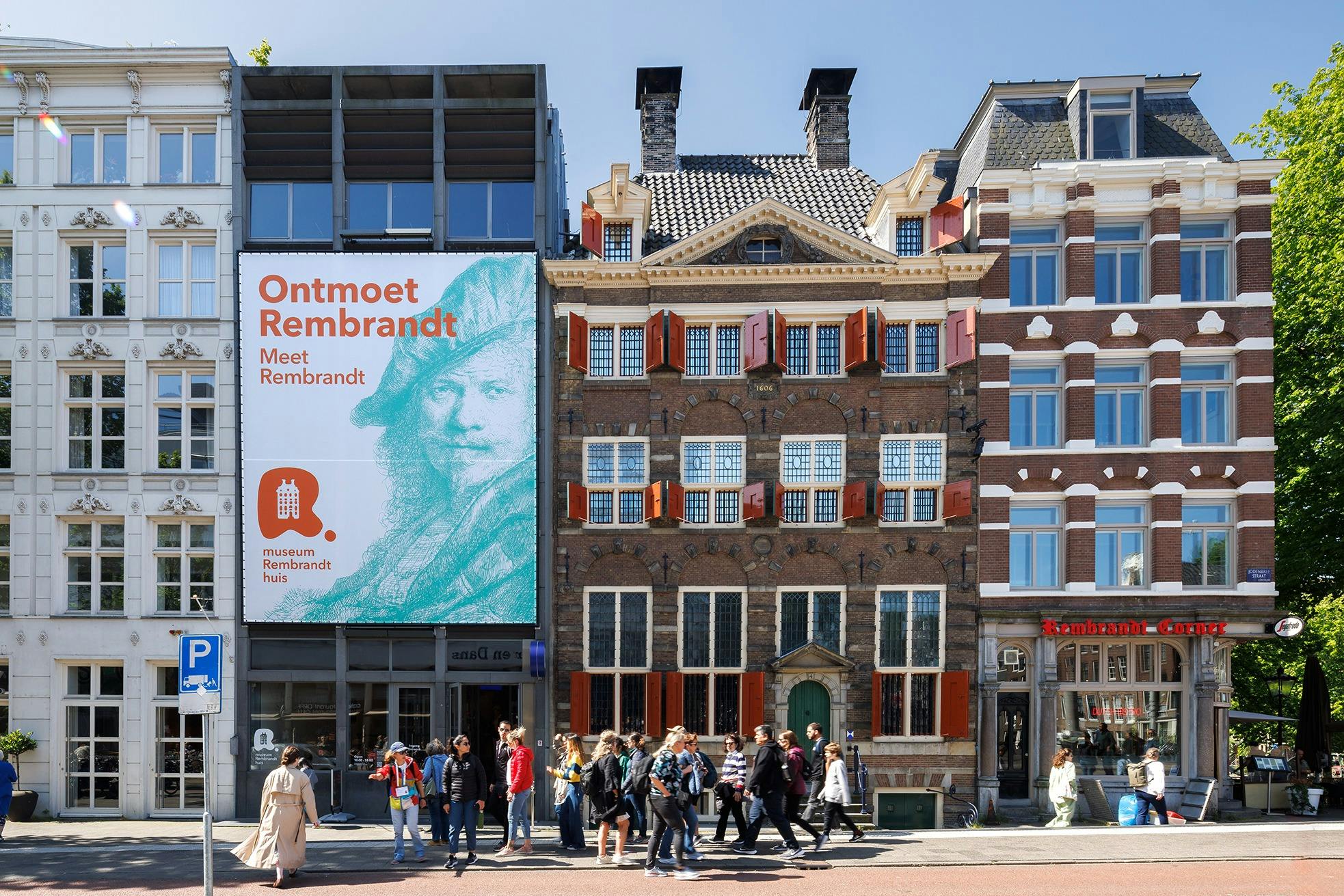 People walk past the Rembrandt House Museum in Amsterdam, with a large "Meet Rembrandt" sign on an adjacent building.