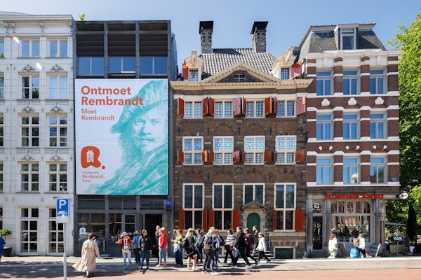 As pessoas passam pelo Museu Casa de Rembrandt em Amesterdão, com um grande cartaz "Conheça Rembrandt" num edifício adjacente.