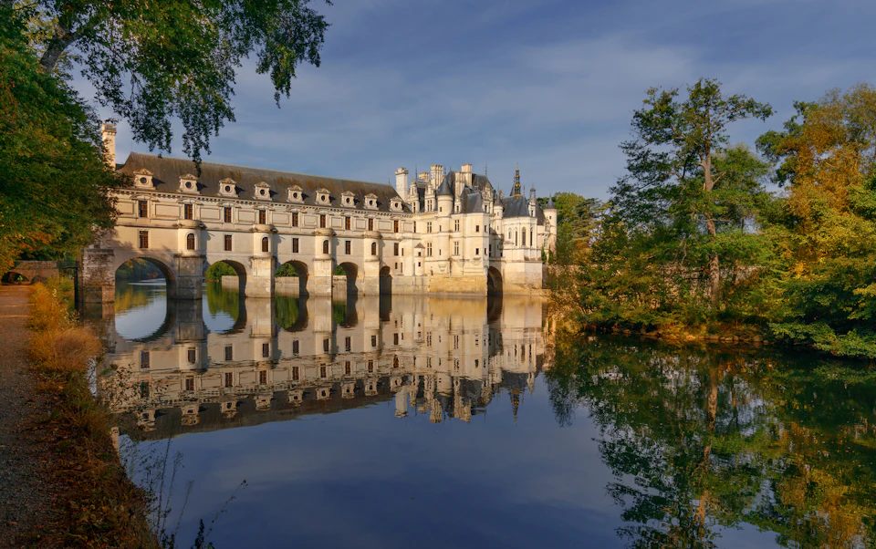 Historic castle with arched bridge over a calm river, reflecting on the water's surface. Surrounded by trees, under a clear sky.