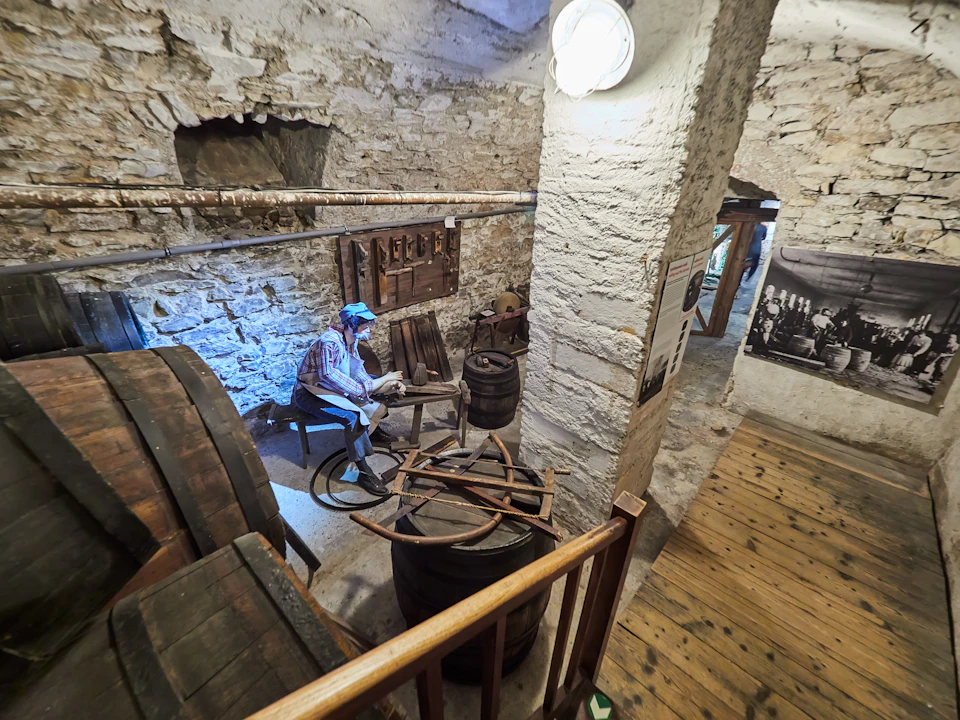 A diorama in a rustic room features a mannequin working with wooden barrels and tools, next to stone walls and a wooden floor.