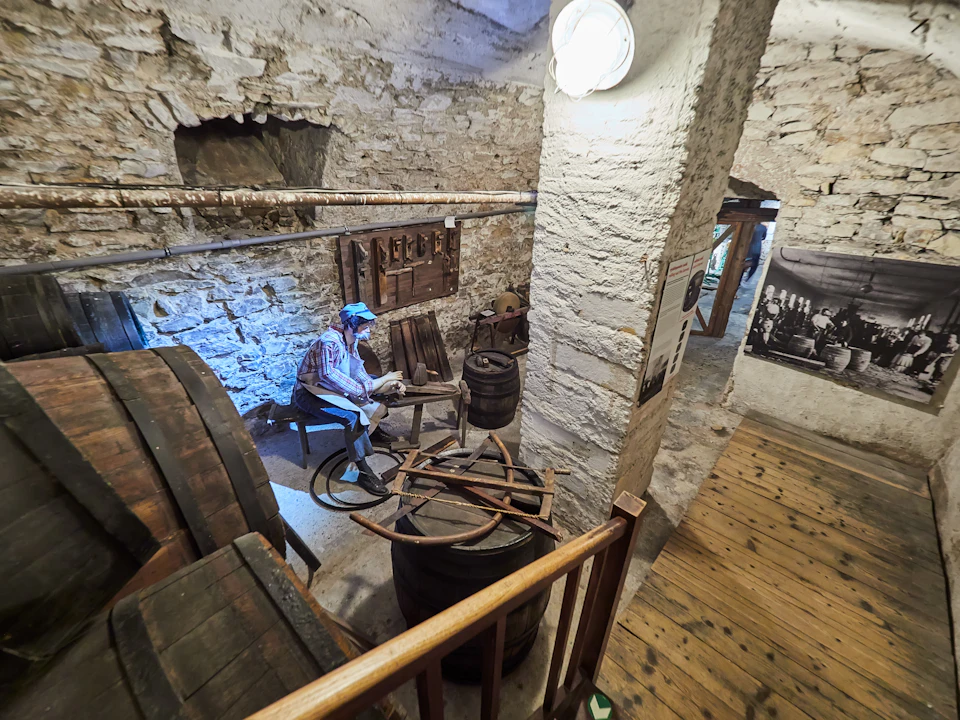 A diorama in a rustic room features a mannequin working with wooden barrels and tools, next to stone walls and a wooden floor.