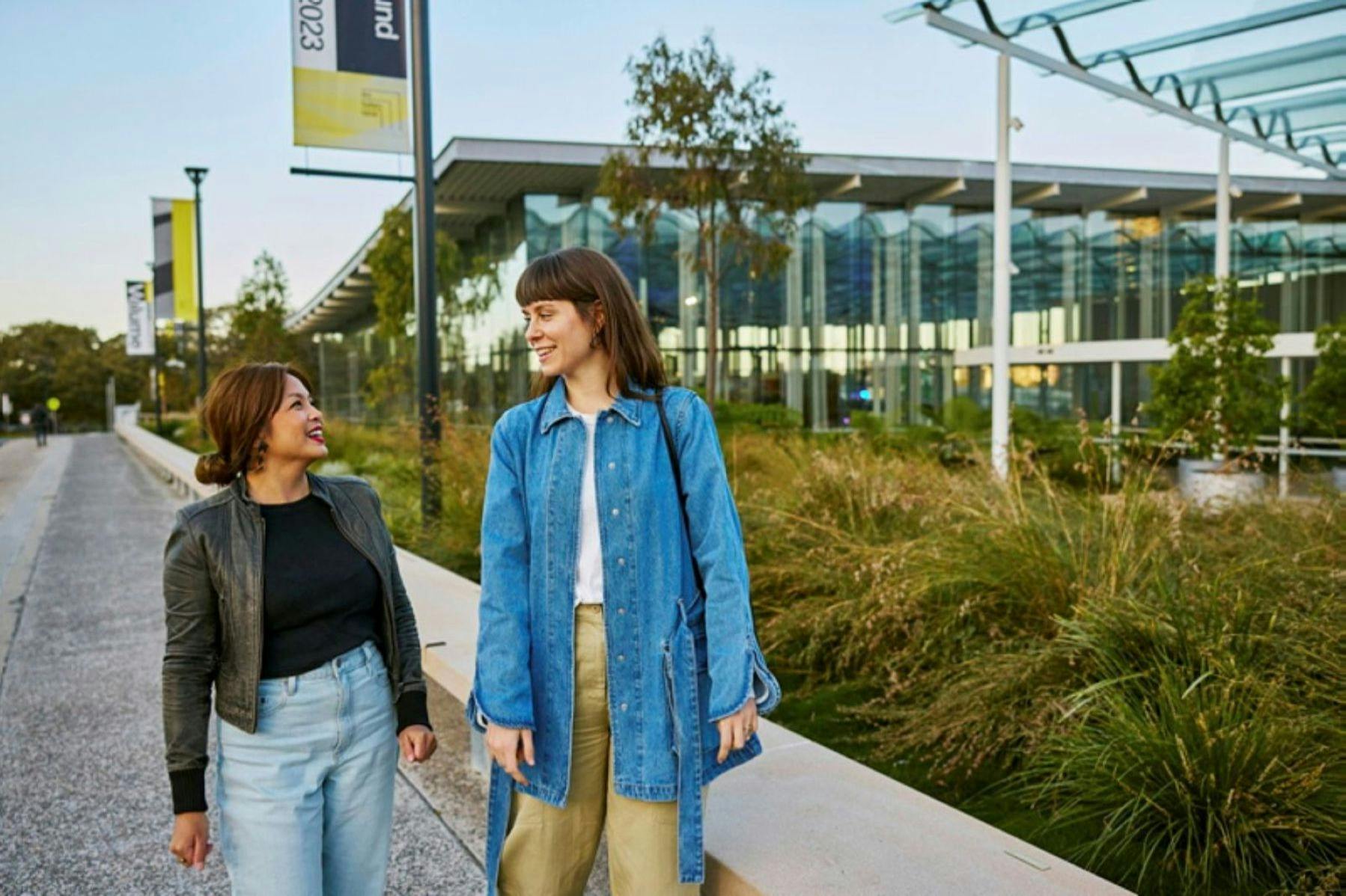 Two women walking and smiling on a pathway next to a modern building and greenery. One wears a denim jacket; the other, a black jacket.