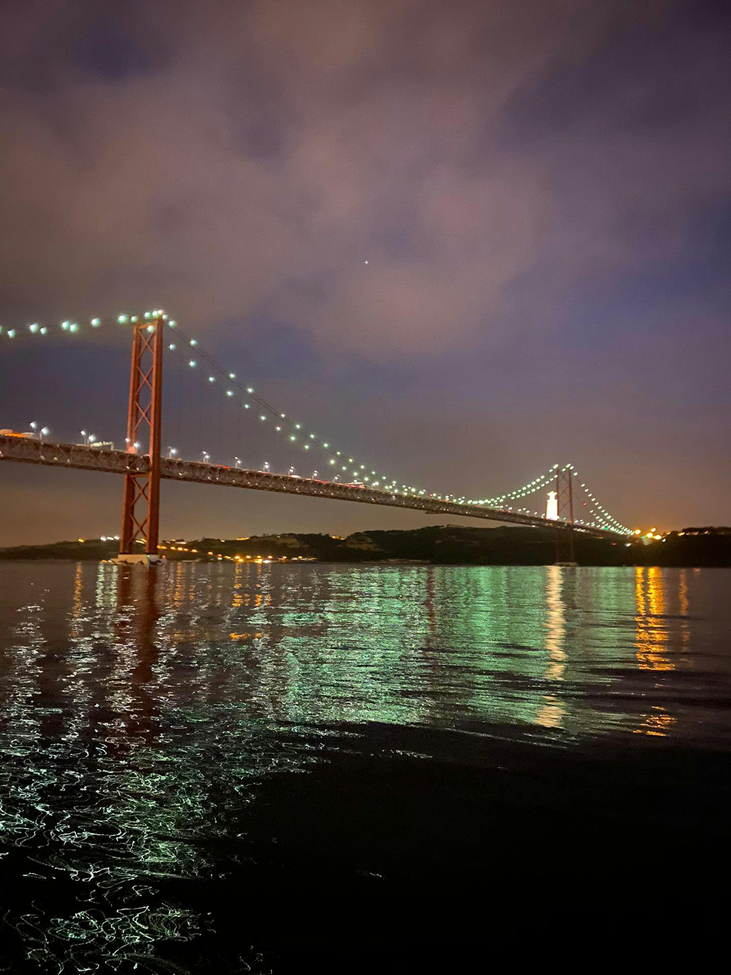 在多云的天空下，一座点燃的吊桥在夜间横跨着一片平静、反光的水域，还有一座遥远的照明雕像。
