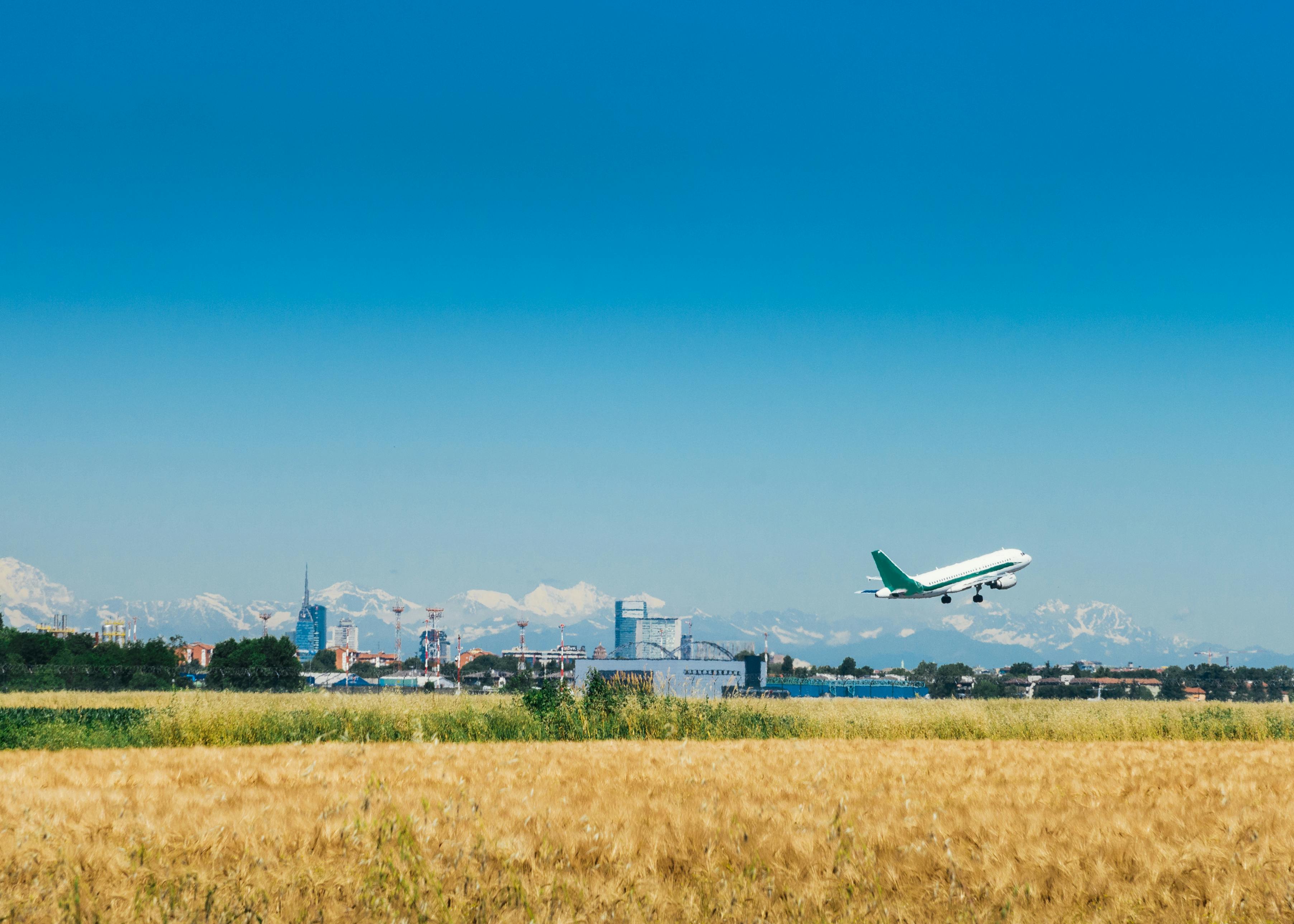 An airplane takes off over a field of wheat, city skyline, and distant snow-capped mountains under a clear blue sky.