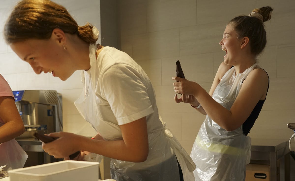 A laughing woman in a plastic apron holds a chocolate dispenser next to another person in a kitchen setting.