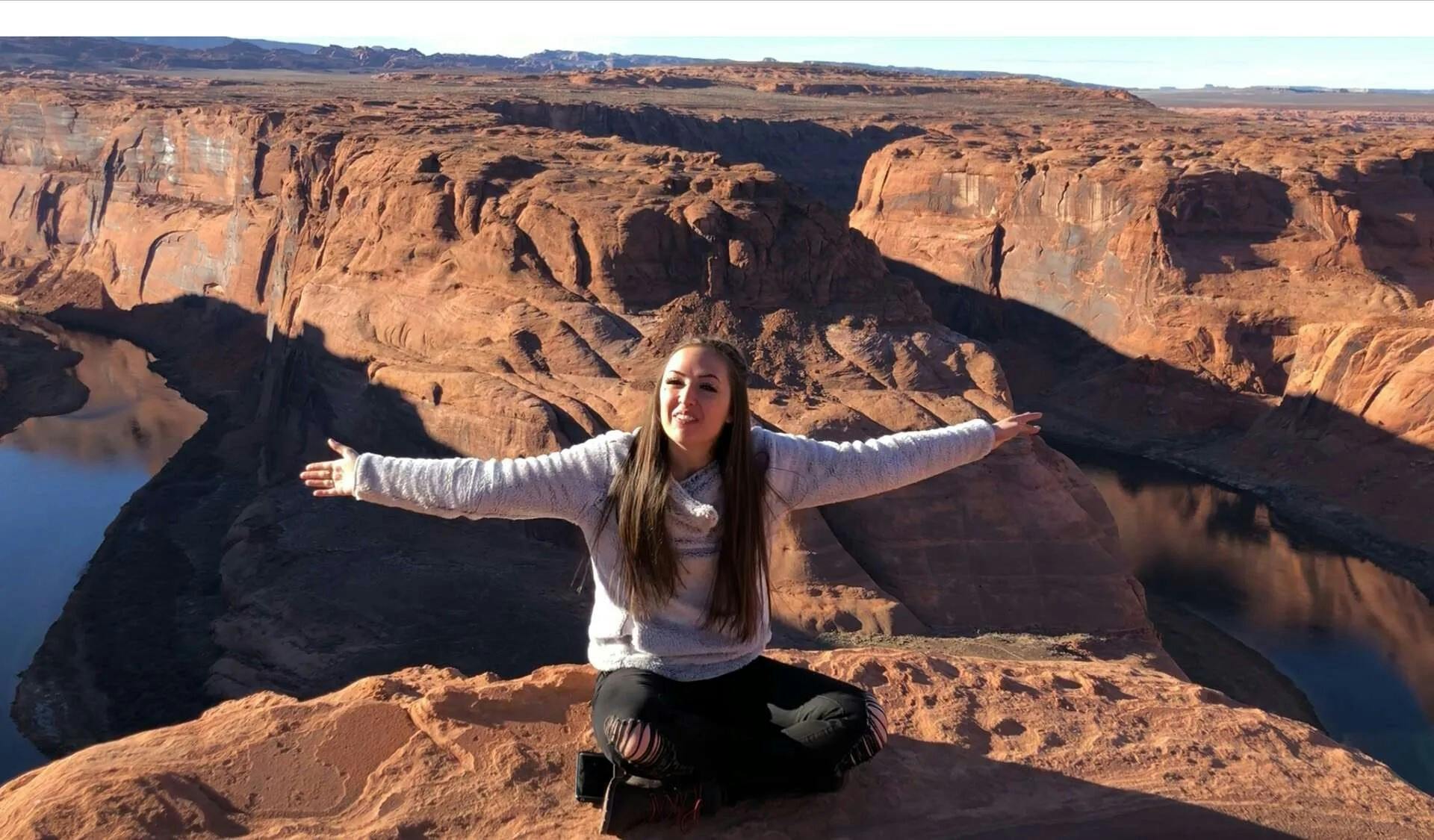 A person sits on a rocky ledge with arms outstretched, smiling, with a vast desert canyon landscape in the background.