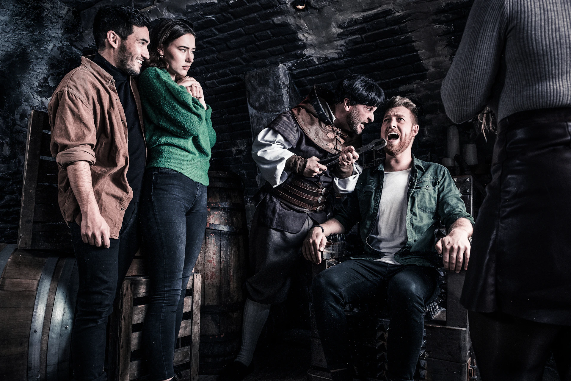 Four people in a dimly lit medieval setting; one is restrained and appears distressed, whilst another is holding a threatening tool. The other two are watching.