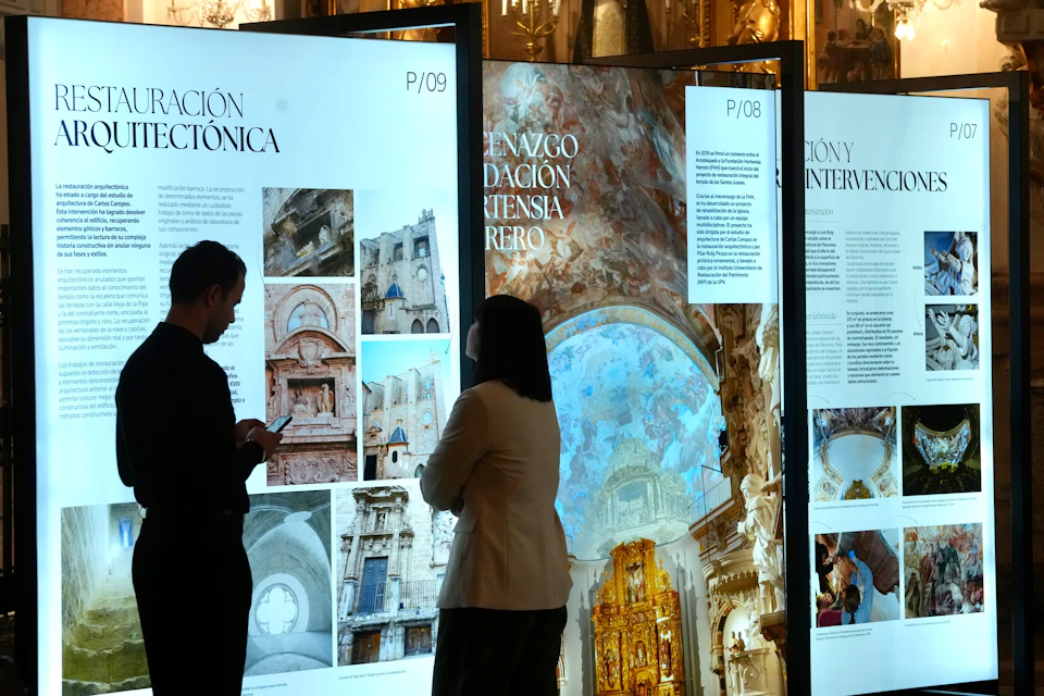 Two people stand in front of illuminated display panels showcasing architectural images and text in an exhibition setting.