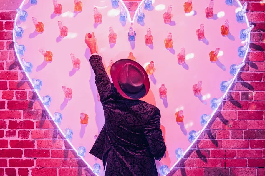 어두운 재킷과 모자를 쓴 사람이 하트 모양의 디스플레이에 담긴 음료수를 찾으려고 손을 뻗습니다. 분홍색으로 불이 켜지고 줄 조명으로 둘러싸여 있습니다.