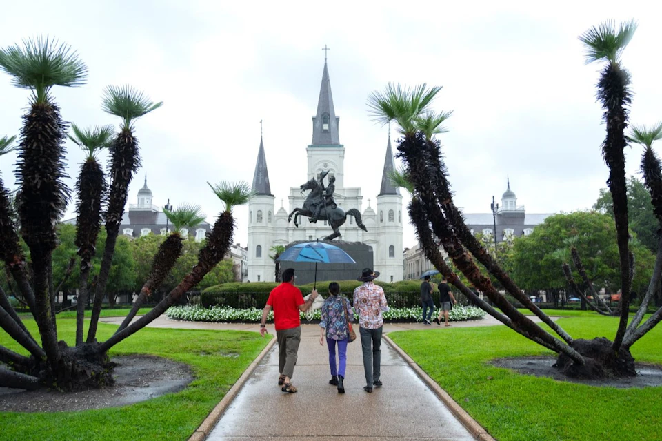 Three people walk towards a white, multi-spired cathedral with a central equestrian statue, surrounded by palm trees and greenery.
